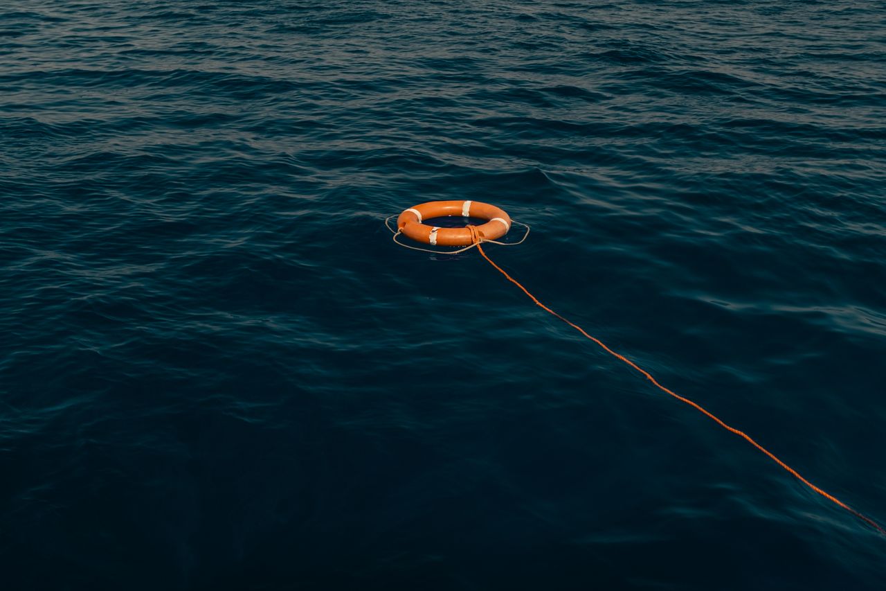 An orange life buoy attached to a rope floating in calm blue waters, symbolizing rescue and safety.